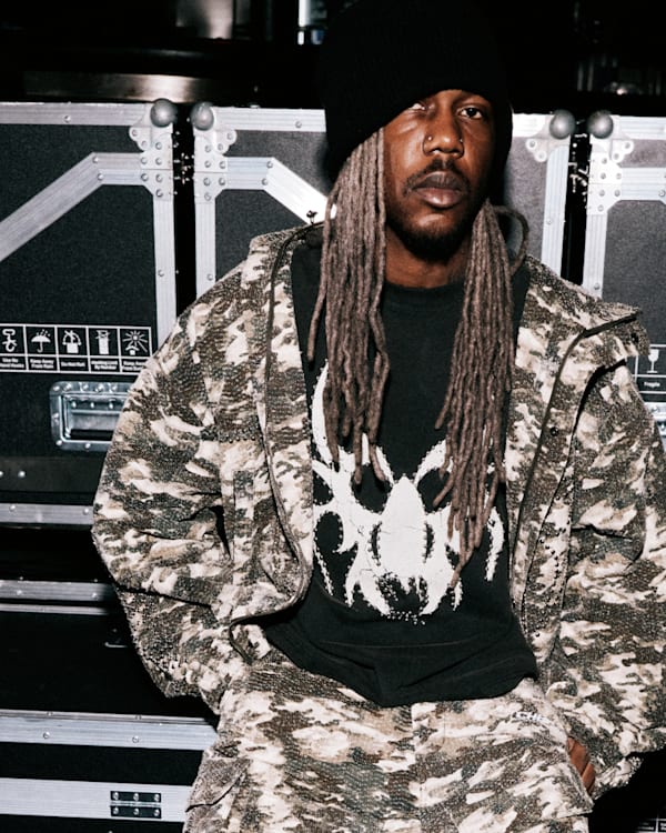 A person with long dreadlocks wearing a patterned outfit and beanie sits in front of stacked equipment cases.