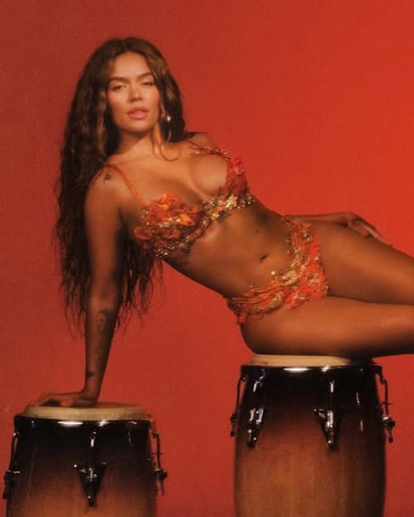 Karol G in a colorful bikini poses on three conga drums against a red background.
