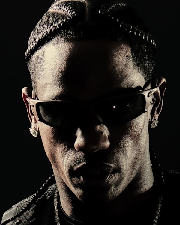 Travis Scott with braided hair and sunglasses, set against a dark background.
