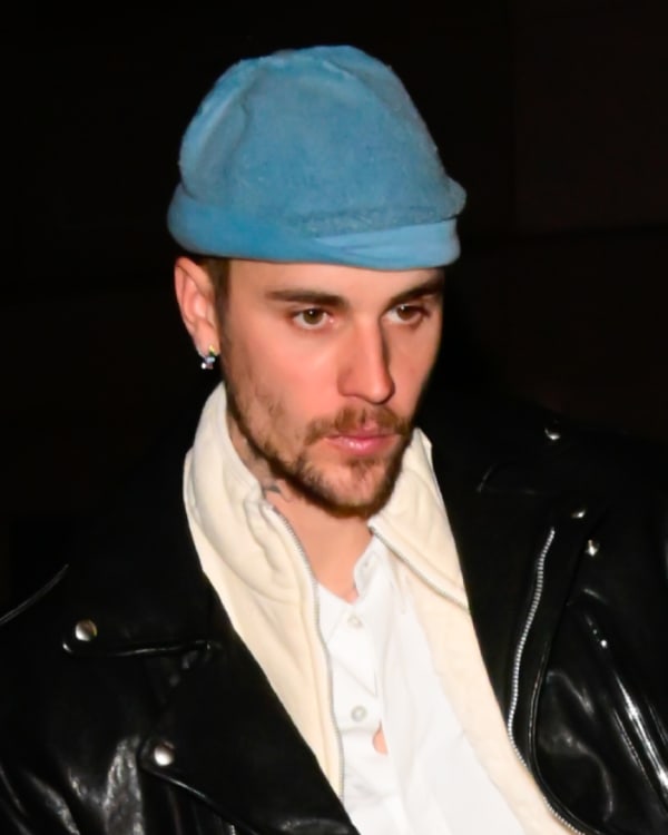 Justin Bieber wearing a blue beanie, black leather jacket, and white shirt, standing outdoors at night.