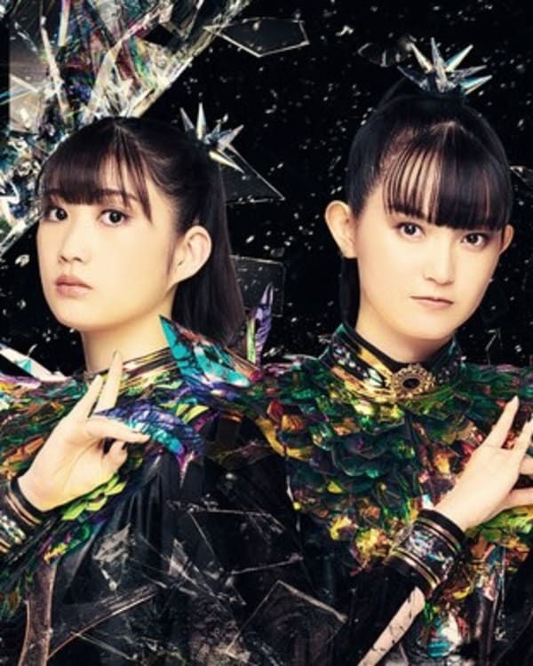 Three members of Babymetal in colorful, futuristic outfits with a dark, abstract background.
