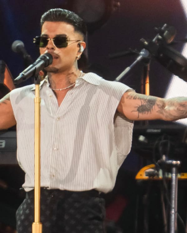 Rauw Alejandro with tattoos and sunglasses performs on stage with arms outstretched, accompanied by a keyboard player in the background.