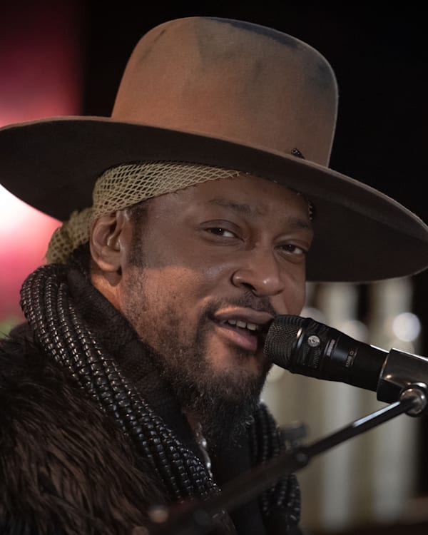 D'Angelo wearing a wide-brimmed hat and scarf sings into a microphone on stage, with a blurred background.