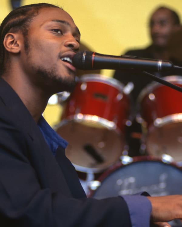A musician with braided hair sings into a microphone while playing the keyboard, with a drummer in the background.