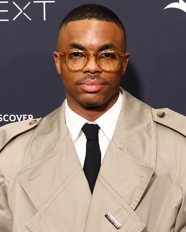 A man in glasses and a beige trench coat stands in front of a branded backdrop.