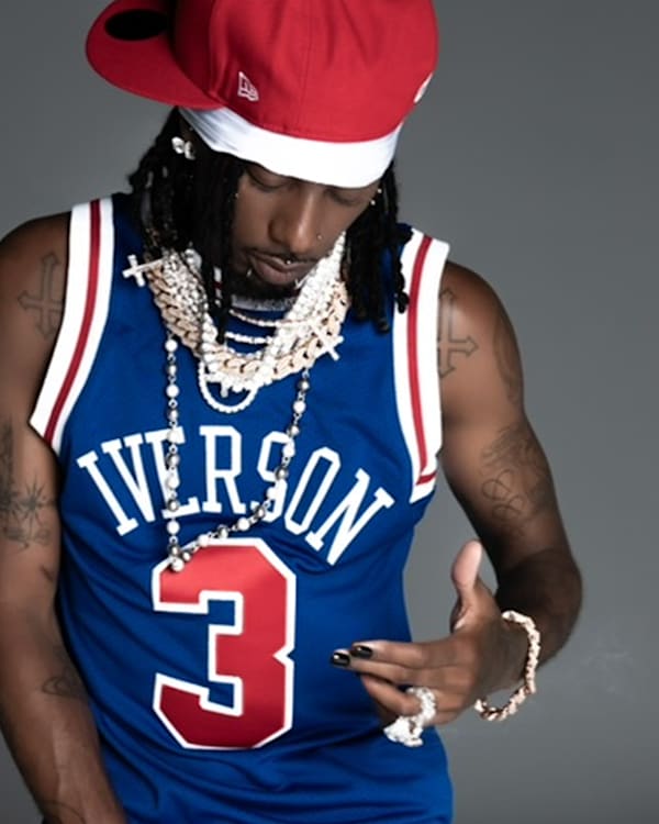 A person wearing an Iverson basketball jersey, red cap, and multiple necklaces, posing with a hand gesture.