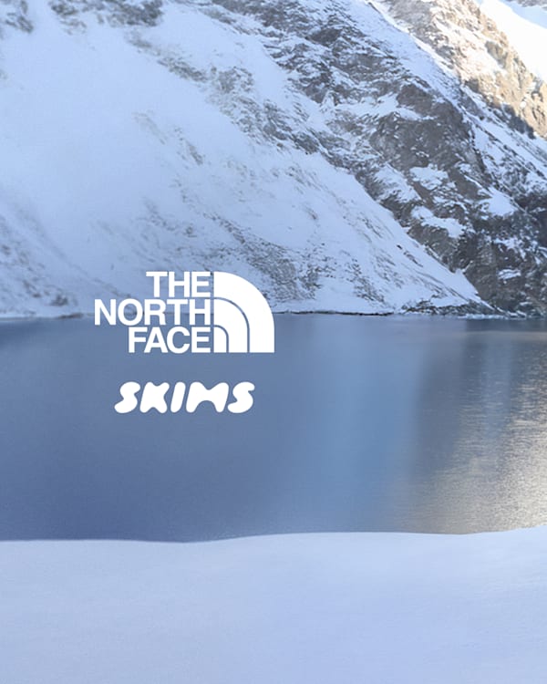 Three people in winter gear from The North Face x Skims collection stand by a snowy lake with mountains in the background.