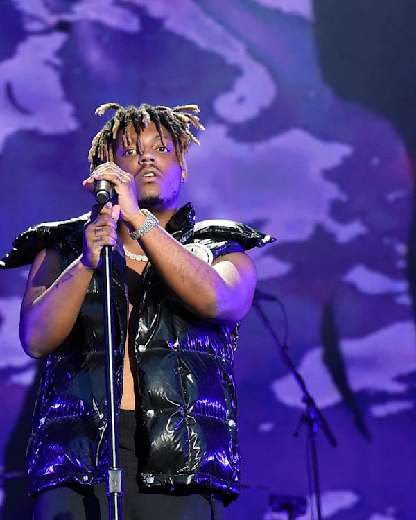 A performer on stage, wearing a shiny black vest, holding a microphone against a purple and blue backdrop.