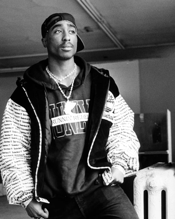 Black and white photo of Tupac Shakur wearing a cap backward, a hoodie, and a jacket, standing in a room with a thoughtful expression.