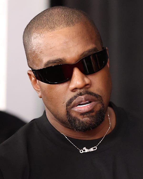 Kanye West wearing sunglasses, dressed in a black shirt, with a serious expression at a public event.
