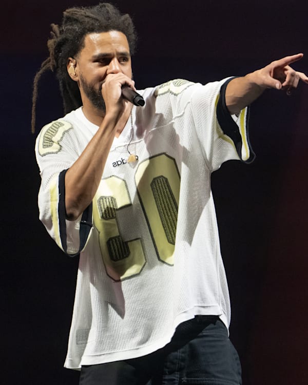 J. Cole with dreadlocks, wearing a white jersey, holds a microphone and points while on stage.