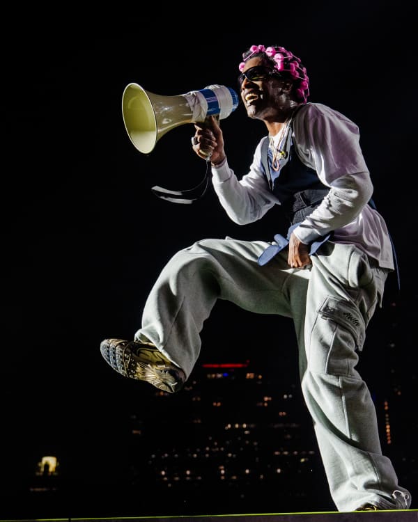 ASAP Rocky with a megaphone, wearing a pink headpiece and casual outfit, energetically poses on stage against a dark cityscape. This performance was in support of his new album, 'Don't Be Dumb.'