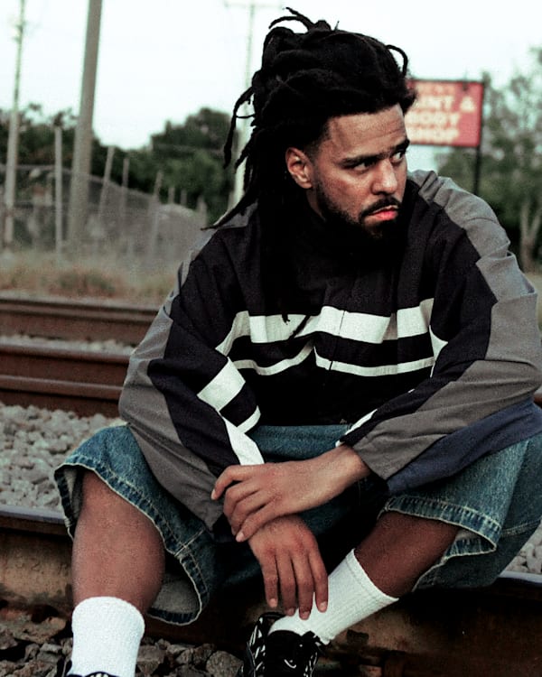 J. Cole with dreadlocks sits on railroad tracks, wearing a striped jacket and denim shorts, looking contemplative.