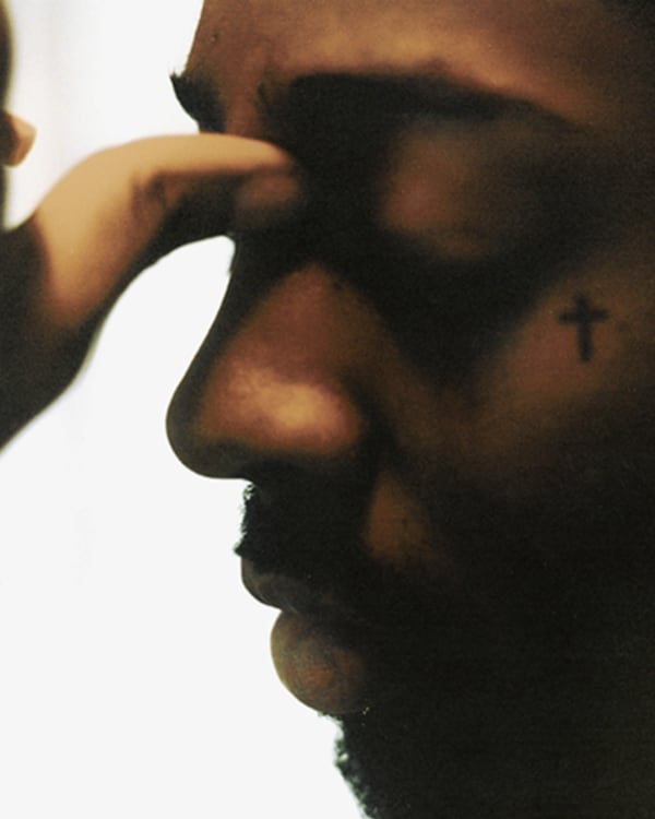 Close-up of a brent fayiaz's face with a cross tattoo near the eye. A hand touches their forehead, and the background is blurred.