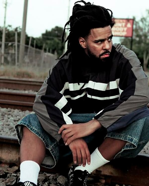 A man with dreadlocks sits on train tracks, wearing a striped jacket and denim shorts, looking thoughtful.