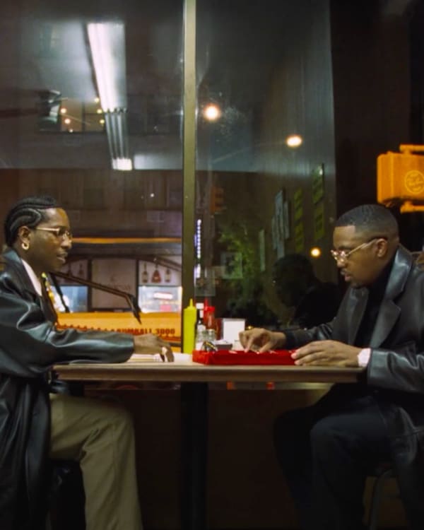 ASAP Rocky and Nas in black leather coats sit across from each other at a diner table with condiments, in a dimly lit urban setting.