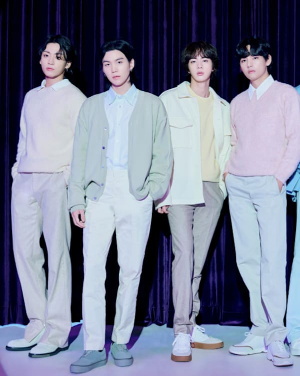 Seven members of BTS stand in a row, wearing pastel-colored outfits, in front of a dark curtain backdrop.