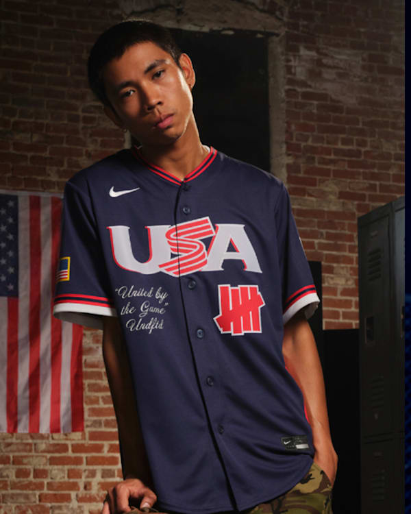 Two men in sports jerseys. The left wears a USA jersey with "Undefeated" branding; the right, a green jersey with "Born x Raised" on the back.