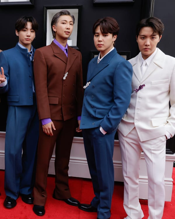 The image shows the members of BTS on a red carpet, each wearing stylish suits in various colors.