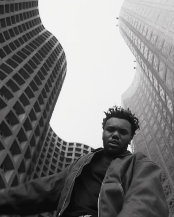 Baby Keem in a jacket is photographed from below between two tall, patterned buildings, creating a dramatic perspective.
