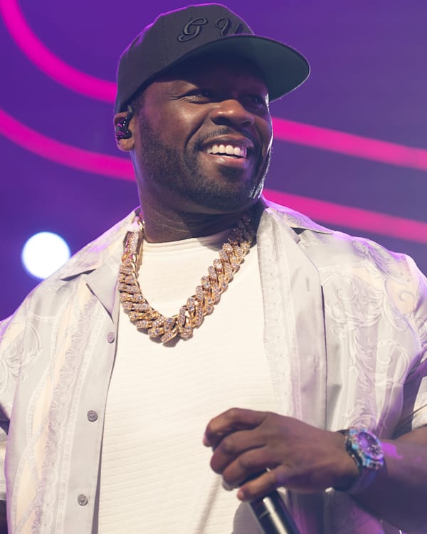 Rapper 50 Cent performing on stage, wearing a cap, white shirt, and gold chain, with colorful lights in the background.