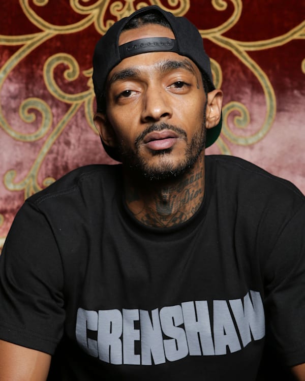 Legendary rapper Nipsey Hussle wearing a black "Crenshaw" shirt and a backward cap sits against a decorative background. Nipsey Hussle was murdered in 2019.