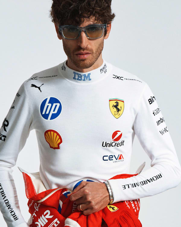 A man wearing Ray-Ban sunglasses and a white Ferrari racing suit with various sponsor logos, holding a red racing jacket. Ray-Ban and Ferrari logos are visible.
