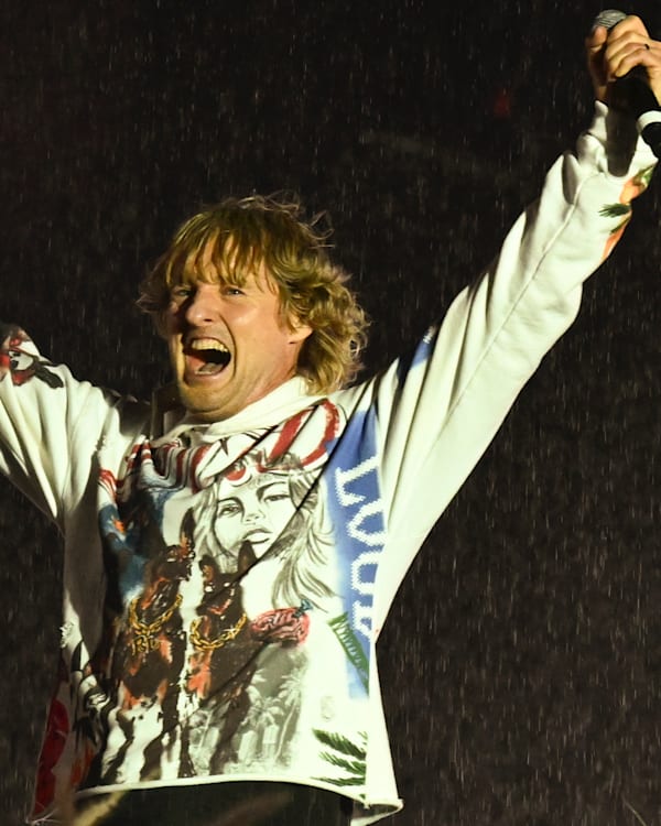Owen Wilson on stage in a graphic sweatshirt, arms raised, holding a microphone, with rain falling around them.