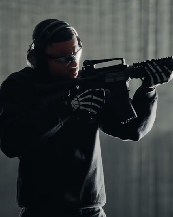 Vince Staples, wearing headphones and skeleton gloves, aims a rifle at the American flag in the video for his song "White Flag."
