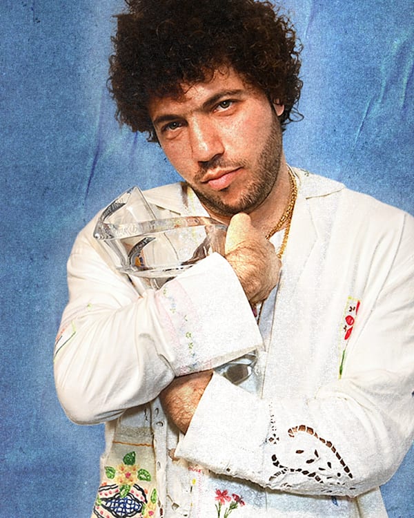Benny Blanco, who has curly hair, in a white embroidered shirt holds a crystal object against a blue textured background.