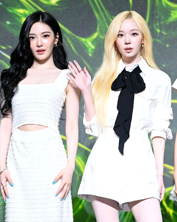aespa's four members stand in a row against a green backdrop, wearing stylish white outfits. They are posing and waving at the camera.