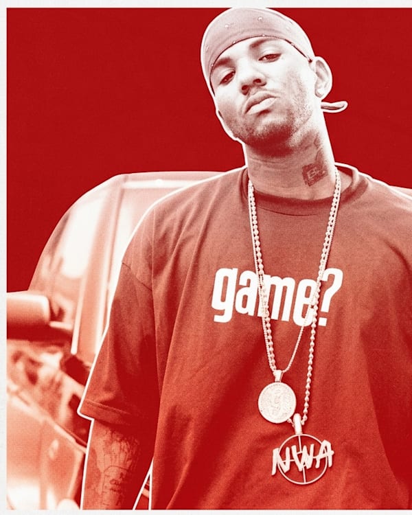 Rapper The Game wearing a bandana and "game?" shirt, standing in front of a car.