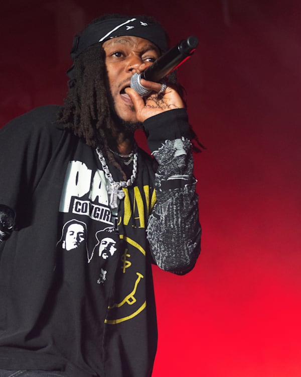 JID with dreadlocks and a black shirt sings into a microphone on stage with red lighting.