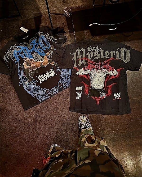 Two WWE t-shirts on the floor: one featuring Rey Mysterio, the other with an RKO design. Person in camo pants and patterned shoes.
