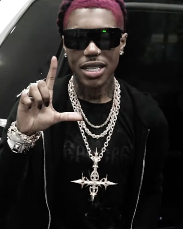 Destroy Lonely with pink hair, sunglasses, and jewelry makes a hand gesture in front of a car.