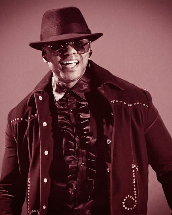 Donell Jones wearing a studded jacket, ruffled shirt, hat, and sunglasses, smiling against a sepia-toned background.
