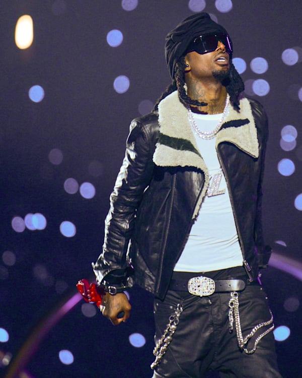 Playboi Carti on stage wearing a black leather jacket and sunglasses, with a backdrop of sparkling lights.