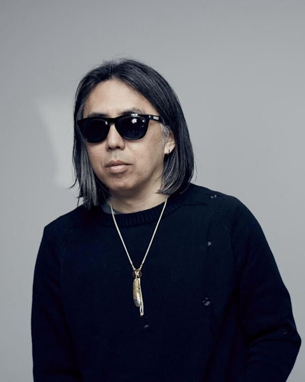 A person with shoulder-length hair, wearing sunglasses, a black shirt, and a necklace, stands against a plain background.