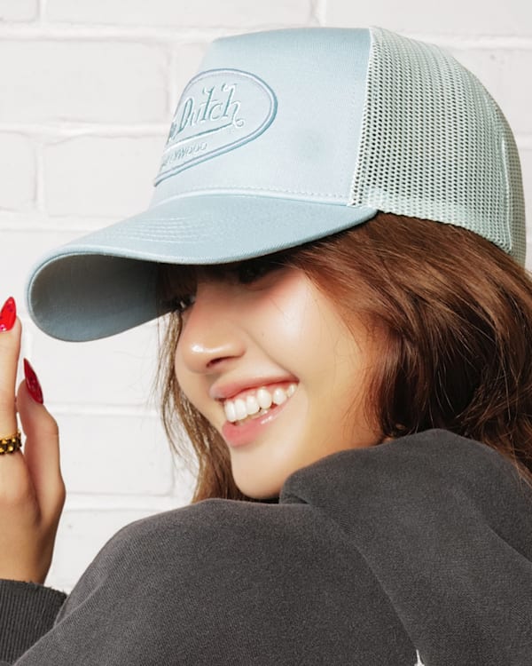 Woman in a light blue cap with red nails, smiling and looking to the side against a white brick wall.