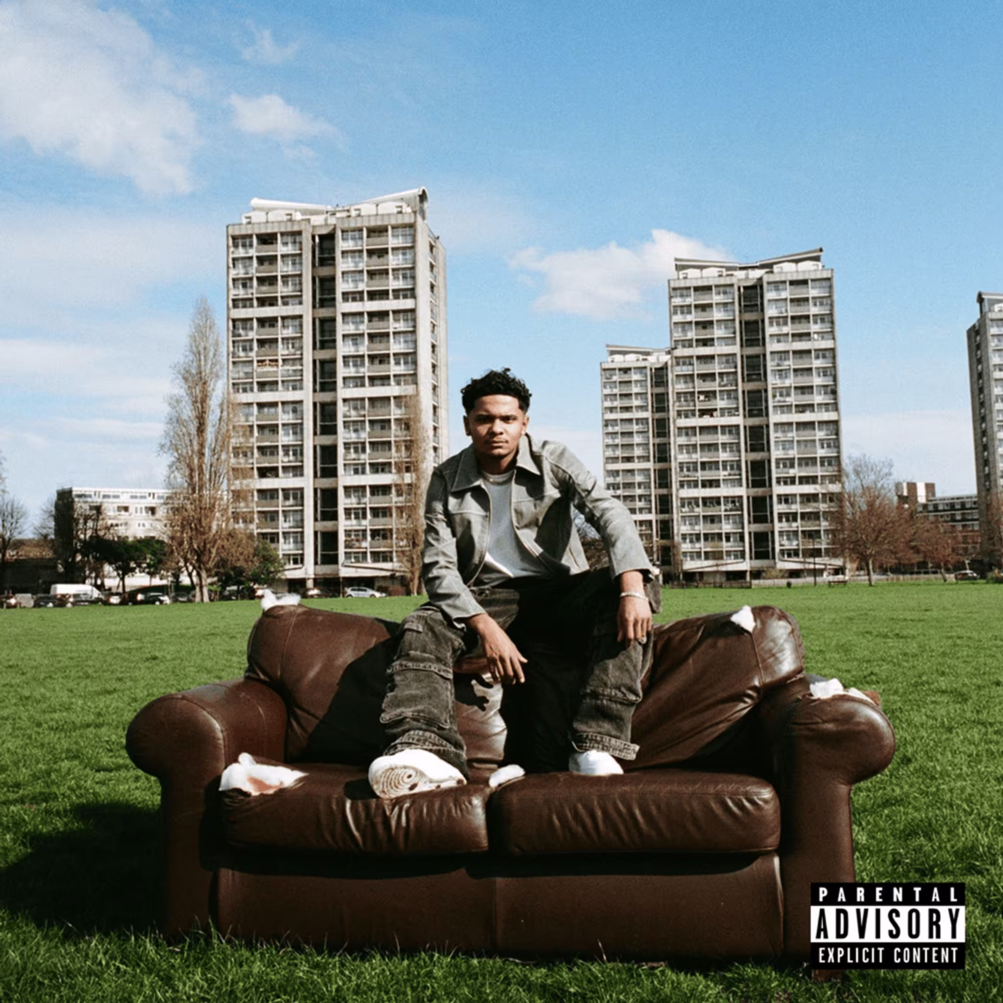 Music artist Roddy Ricch sits on a brown couch in a grassy field with tall apartment buildings in the background. Parental Advisory label in the bottom right