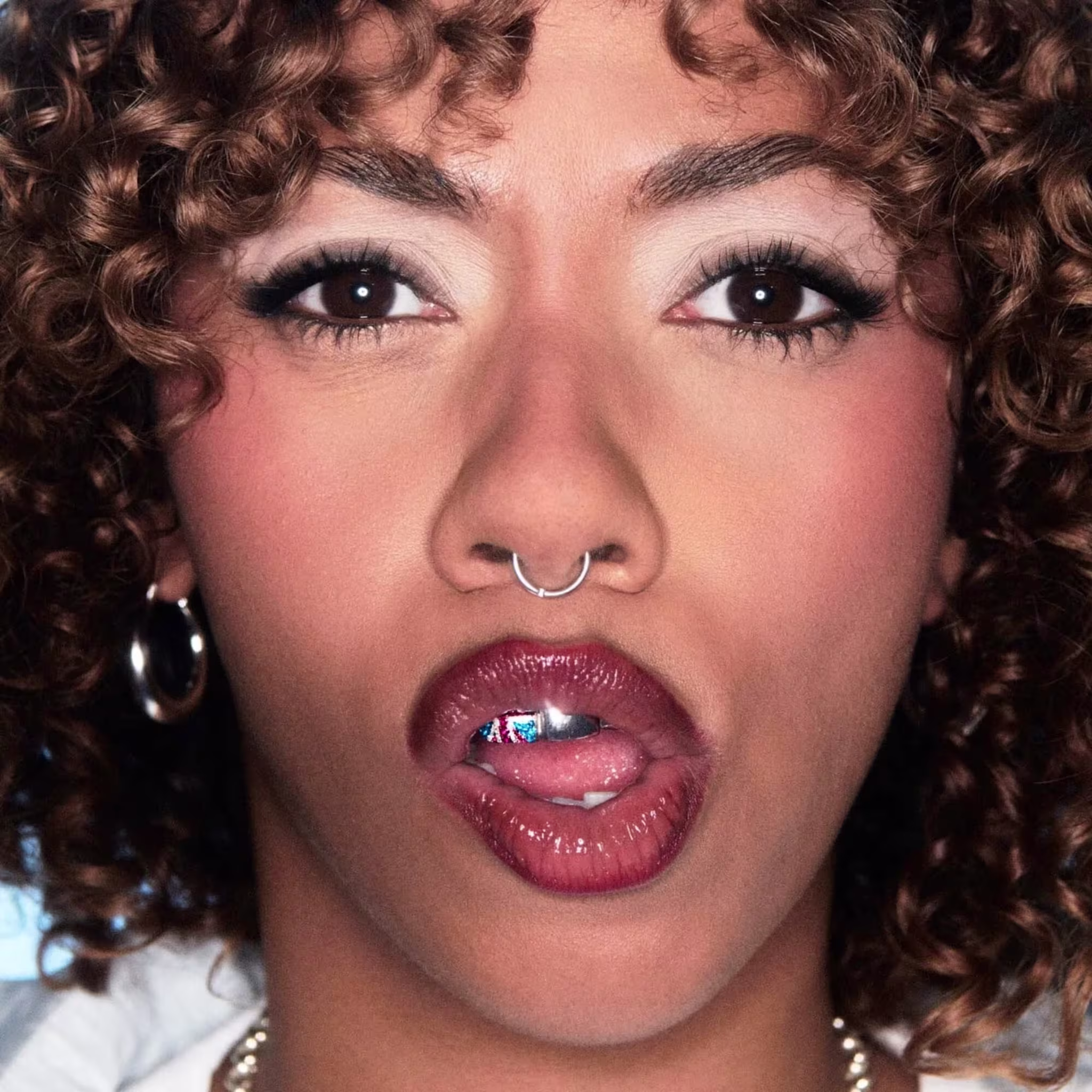Close-up of a person with curly hair, nose ring, and a jewel between their teeth
