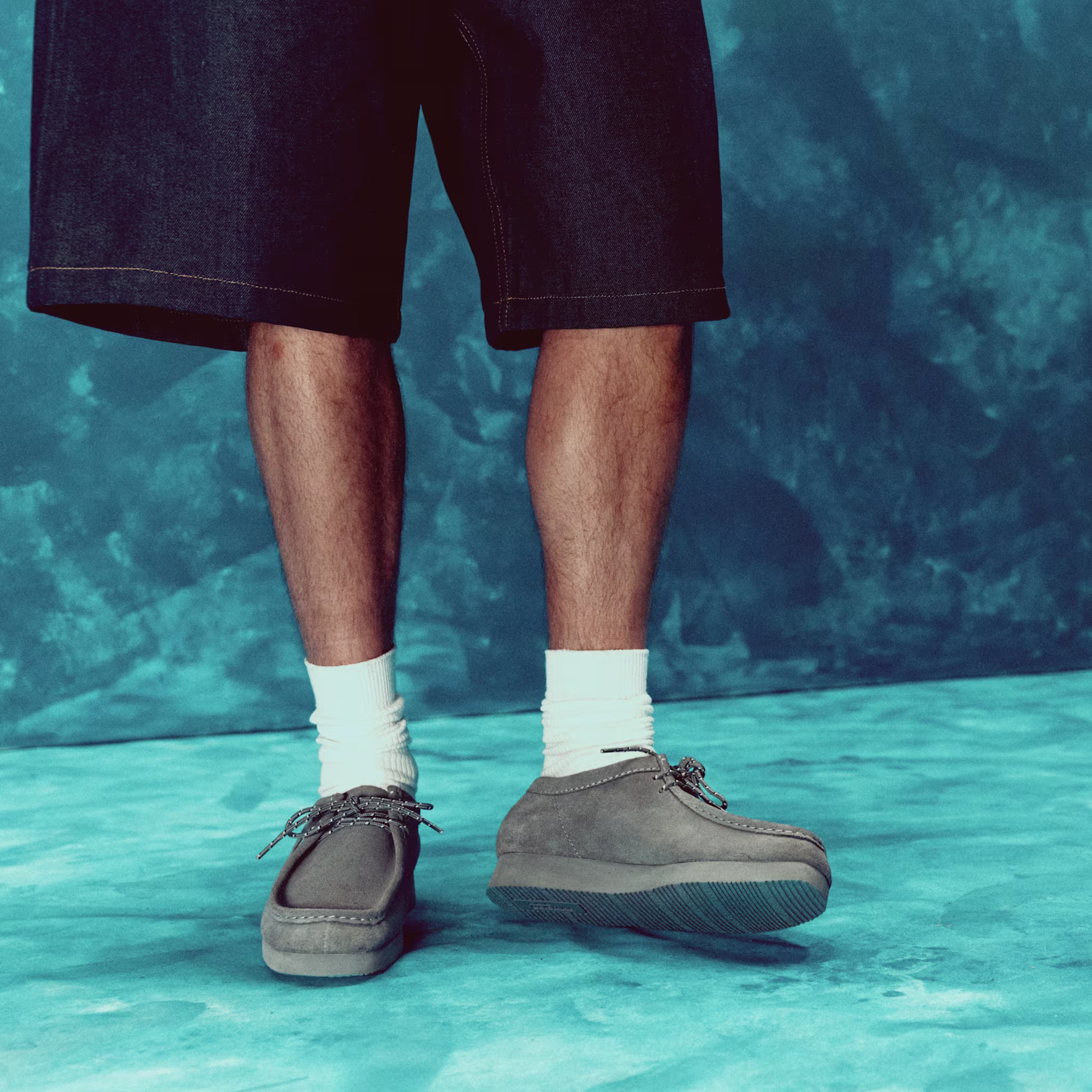 Man wearing denim shorts, q socks, and gray shoes stands on a blue textured floor.