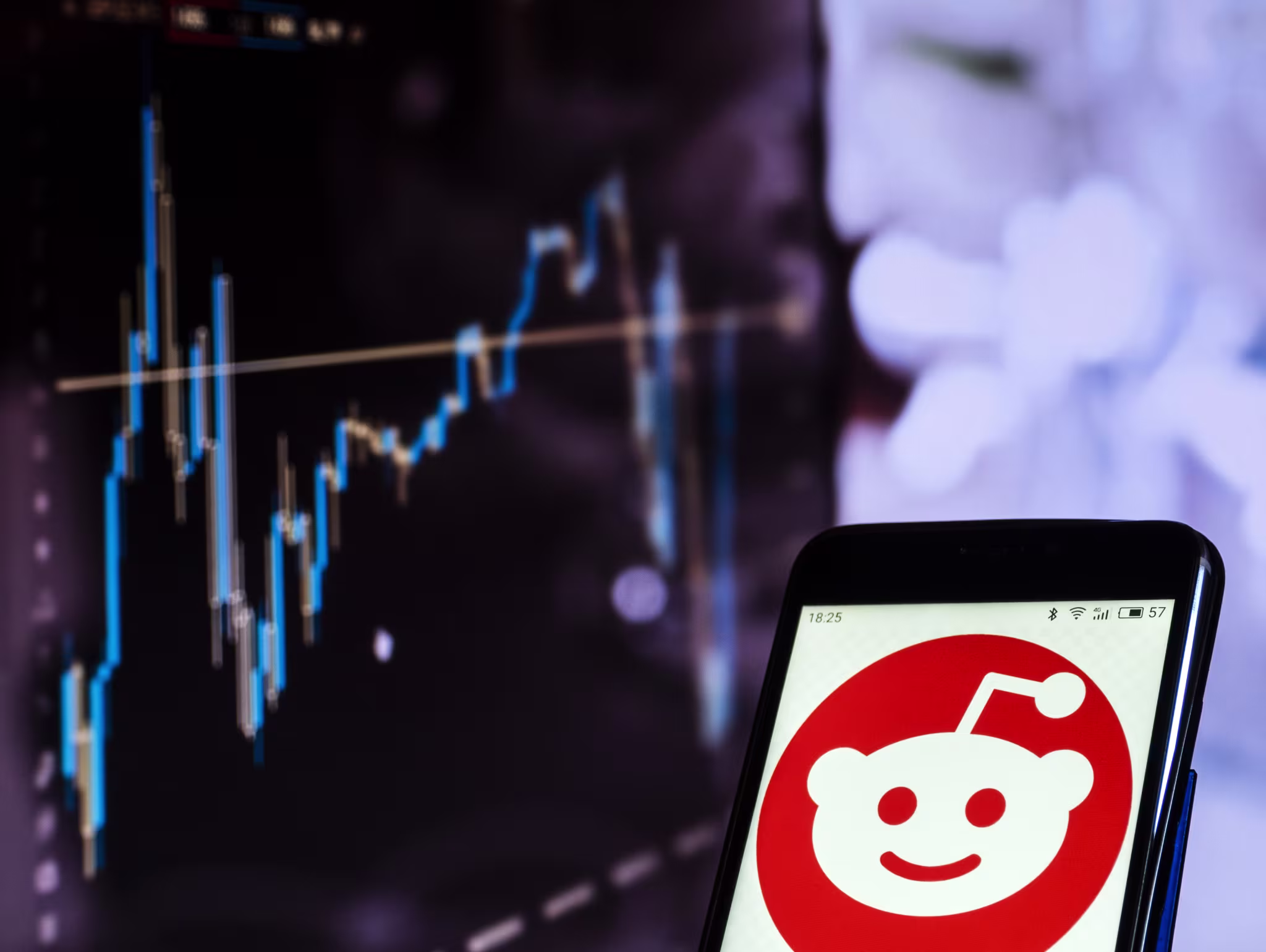 The Reddit logo on a phone screen, in front of a computer screen with a graph on it.