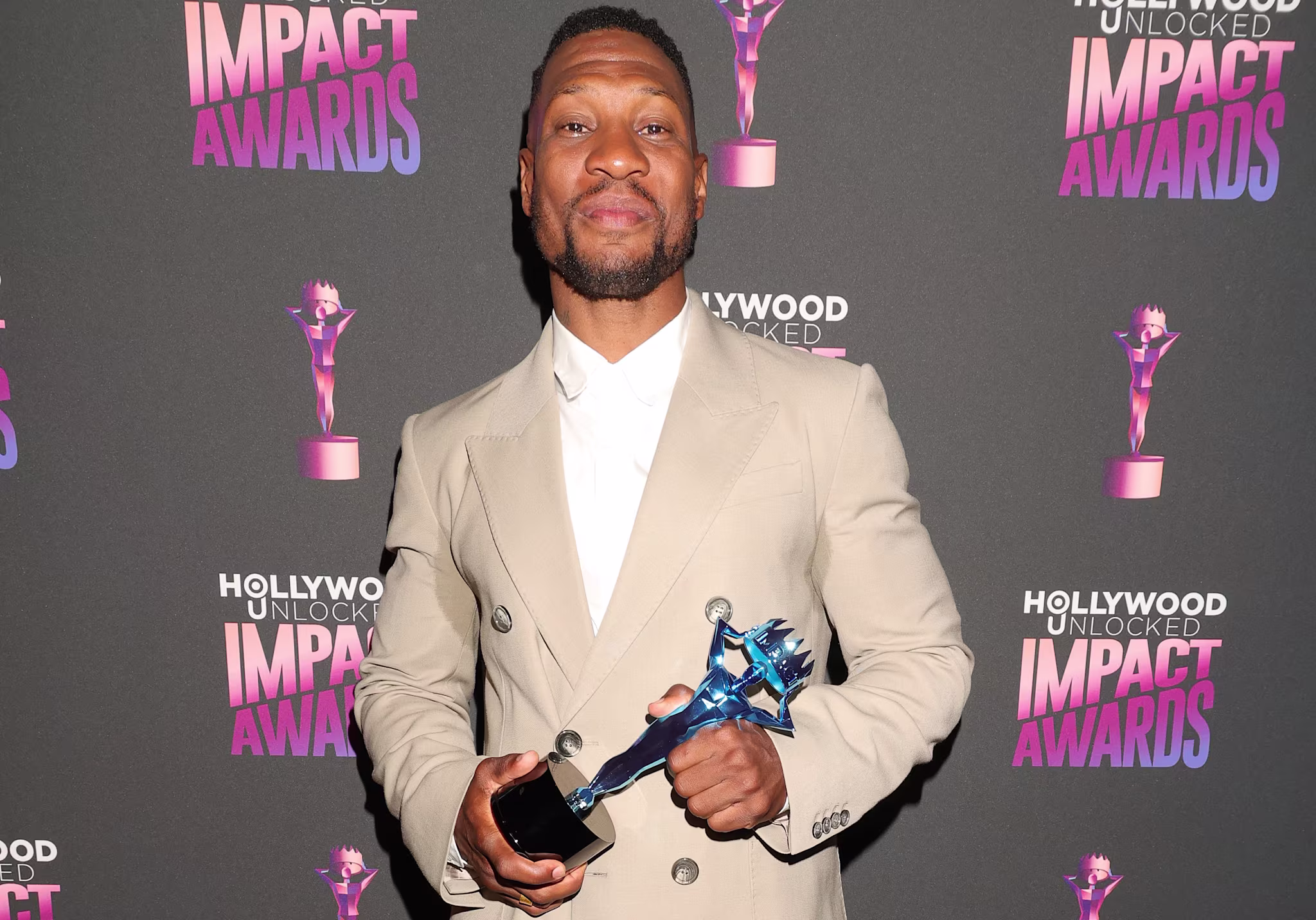 Man in tan suit holding a trophy, posing on the red carpet at the Hollywood Unlocked Impact Awards. Background features event logos repeated