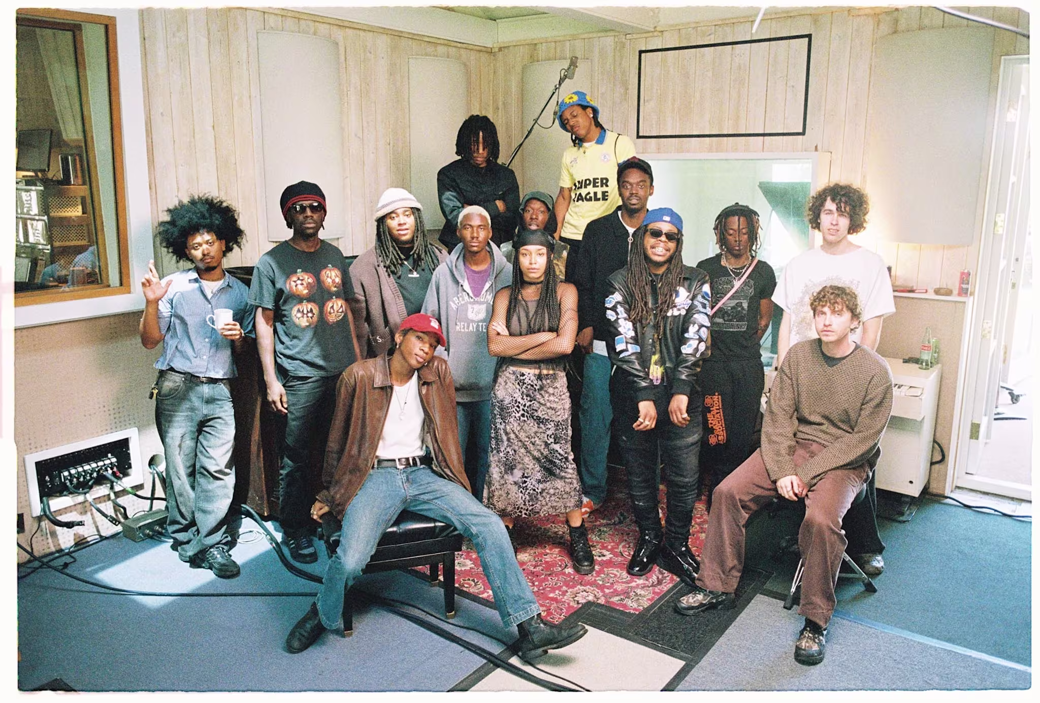 A group of 13 people posing in a recording studio, with casual attire and diverse hairstyles, standing and sitting together.