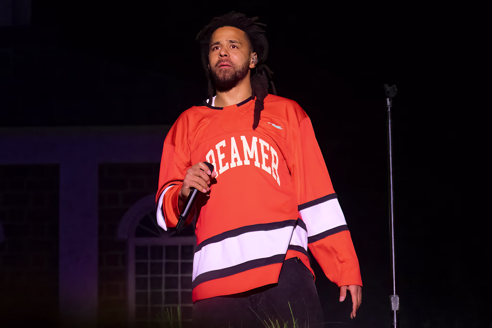 J. Cole performing on stage wearing a "Dreamer" hockey jersey, holding a microphone