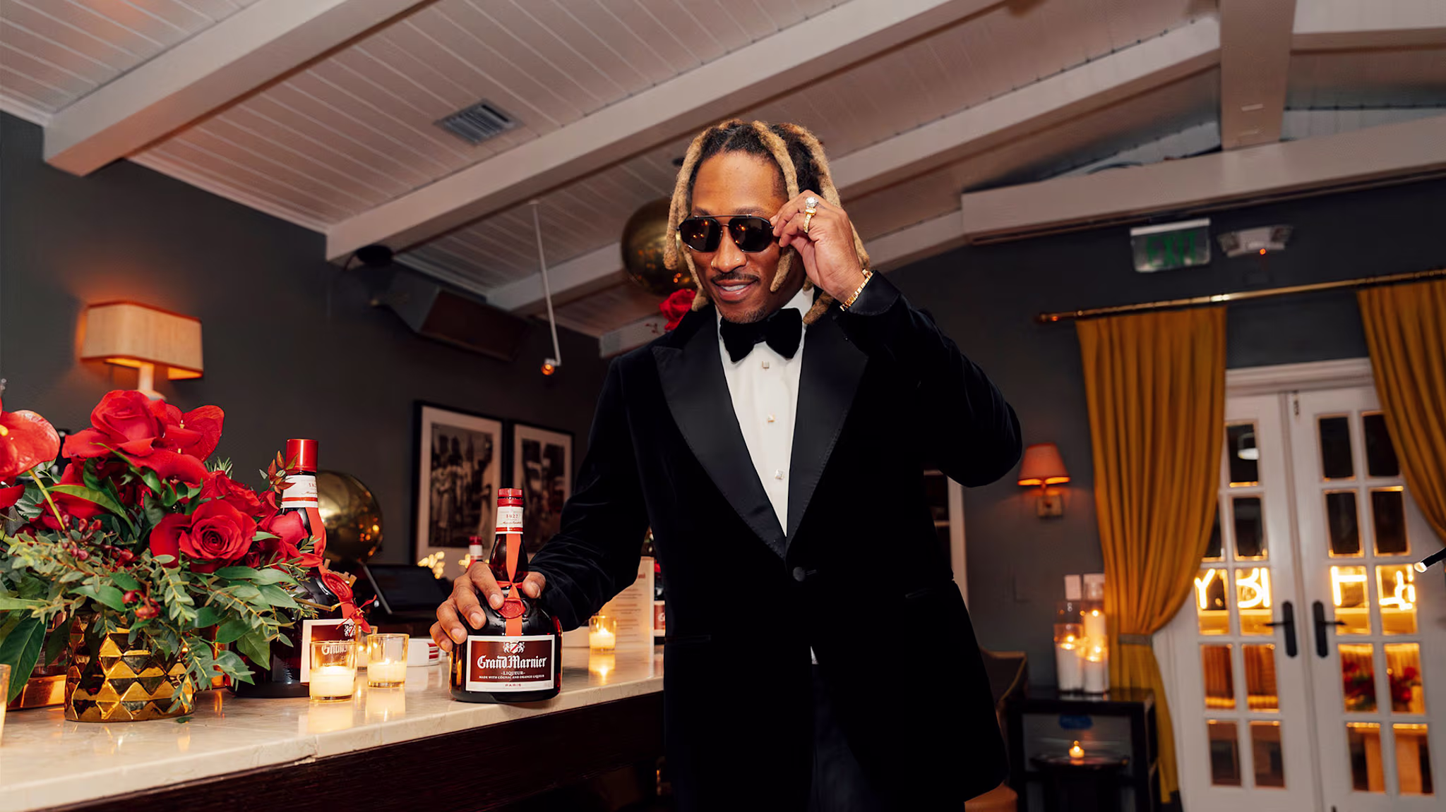 A man in a tuxedo and sunglasses stands at a bar holding a bottle of Grand Marnier, with red flowers and lit candles nearby.