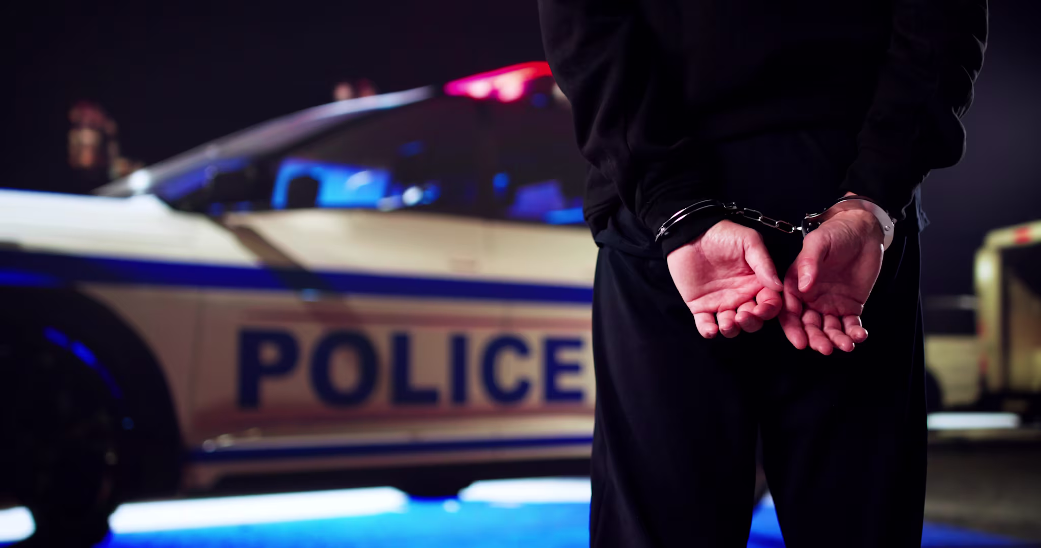 A person in handcuffs stands in front of a police car at night.