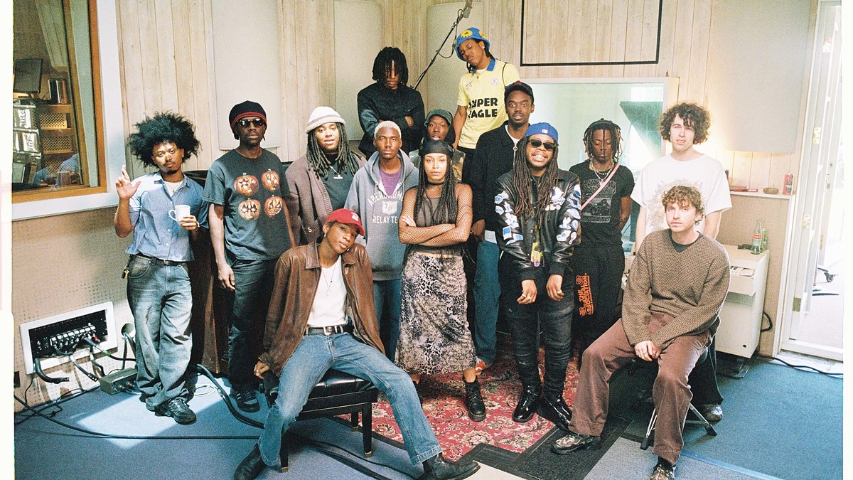 A group of thirteen people in a recording studio, wearing casual and eclectic outfits, standing and sitting together, with musical equipment in the background.