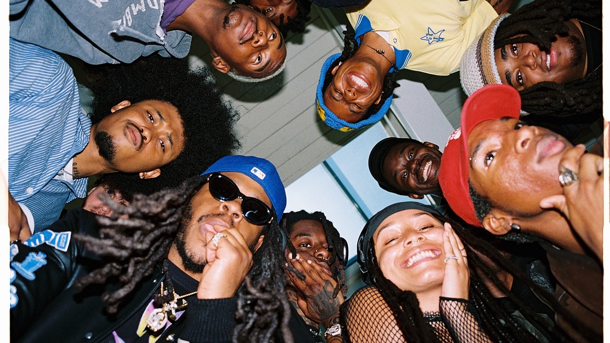 A group of people gathered in a circle, looking down at the camera, smiling and making playful expressions, with varied hairstyles and hats.